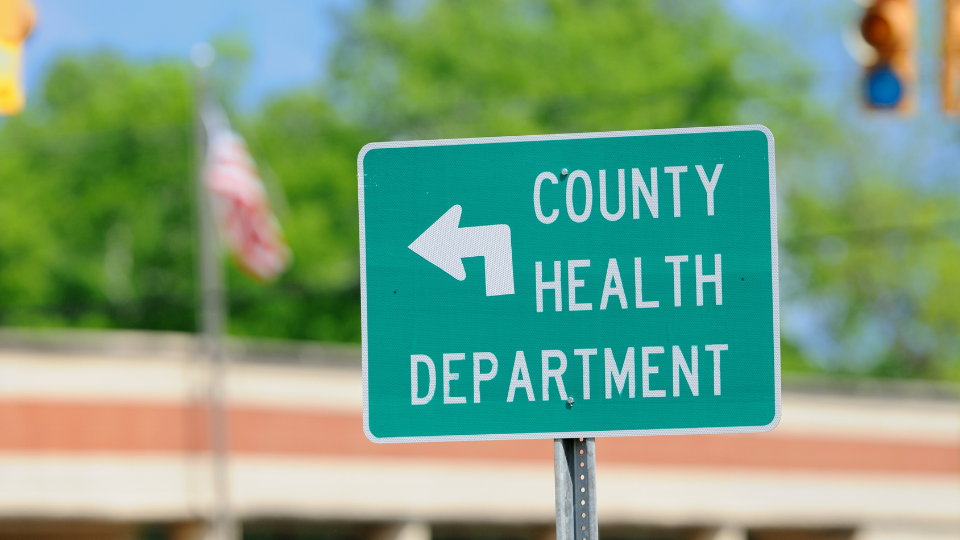 A green County Health Department sign with an American flag in the background